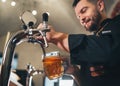 Tapping fresh lager beer in glass mug close up. Smiling stylish bearded barman dressed black uniform with an apron at bar counter Royalty Free Stock Photo