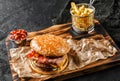 Tasty burger with beef meat cutlet, slice of cheese, ham and spicy sauce on wooden board with french fries potatoes on black Royalty Free Stock Photo