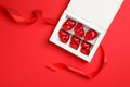 Tasty chocolate heart shaped candies in white box with ribbon on red background, flat lay Royalty Free Stock Photo