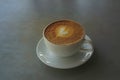 Tasty drinking, a cup of capuccino coffee decorated with heart pattern on brown milk froth in white ceramic cup on gray table Royalty Free Stock Photo