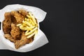 Tasty fastfood: fried chicken drumsticks, spicy wings, French fries and chicken fingers in paper box over black background, top vi