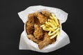 Tasty fastfood: fried chicken legs, spicy wings, French fries and chicken fingers in paper box over black background, overhead vie