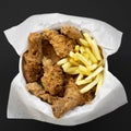 Tasty fastfood: fried chicken legs, spicy wings, French fries and chicken fingers in paper box over dark black background, top