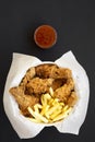 Tasty fastfood: fried chicken legs, spicy wings, French fries and chicken tenders in paper box, sour-sweet sauce on black surface