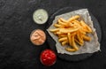 Tasty french fries potatoes on paper with sauces over black stone background. Hot fast food, top view Royalty Free Stock Photo