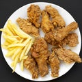Tasty fried chicken drumsticks, spicy wings, French fries, chicken fingers on white plate over black surface, top view. Flat lay, Royalty Free Stock Photo