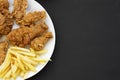 Tasty fried chicken drumsticks, spicy wings, French fries, chicken fingers on white plate over black surface, top view. Flat lay, Royalty Free Stock Photo
