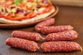 Tasty little sausages salami on brown wooden table close up. On blurred background pizza with tomatoes, bell pepper, onion Royalty Free Stock Photo