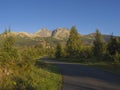 Tatra Mountains range with asphalt road curve, golden hour light, way from Tatranska Lomnice to Start with green trees and grass.