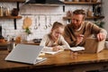 Tattooed and bearded father holding book while daughter doing homework near laptop in kitchen at home Royalty Free Stock Photo