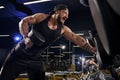 Tattooed, bearded muscular man in black shorts, vest, cap. Exercising with dumbbells for training his triceps Dark gym Royalty Free Stock Photo