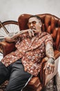 Tattooed gangster yawns while sitting on a luxurious retro armchair, looks very expressive and freakly