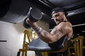 Tattooed guy with earring, in black vest and cap. He is lifting a dumbbell, training his biceps, sitting on preacher Royalty Free Stock Photo