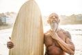 Tattooed senior surfer holding vintage surf board on the beach at sunset - Focus on face