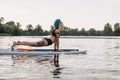 tattooed woman with blue hair practicing yoga on paddleboard in water. Upward facing dog pose