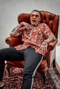 Tattooed gangster yawns while sitting on a luxurious retro armchair, looks very expressive and freakly