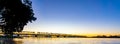 Tauranga`s historic railway bridge silhouetted by golden sunrise across harbour with pohutukawa tree in foreground