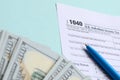 1040 tax form lies near hundred dollar bills and blue pen on a light blue background. US Individual income tax return