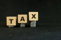 Tax increase concept. Increasing stack of coins in wooden blocks on dark black background.