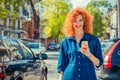 Taxi app. Woman using smart phone, standing on city street on sunny day waiting for taxi. Beautiful redhead girl holding mobile, Royalty Free Stock Photo