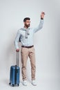 Taxi. Young and handsome man with a luggage is raising his hand while standing in studio on a grey background. Full Royalty Free Stock Photo