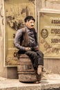 Figurine of wrinkled old mustached man in leather vest and cap sitting on a wine barrel holding a bottle