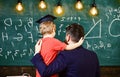 Teacher with beard, father hugs little son in classroom while discussing, chalkboard on background. Child in graduate Royalty Free Stock Photo