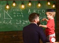 Teacher with beard, father teaches little son in classroom, chalkboard on background. Boy, child in graduate cap Royalty Free Stock Photo