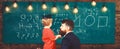 Teacher with beard, father teaches little son in classroom, chalkboard on background. Boy, child in graduate cap