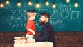 Teacher with beard, father teaches little son in classroom, chalkboard on background. Boy, child in graduate cap play Royalty Free Stock Photo