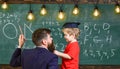 Teacher with beard, father teaches little son in classroom, chalkboard on background. Child in graduate cap listening