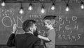 Teacher with beard, father teaches little son in classroom, chalkboard on background. Child in graduate cap listening