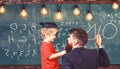 Teacher with beard, father teaches little son in classroom, chalkboard on background. Child in graduate cap listening Royalty Free Stock Photo