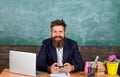 Teacher bearded hipster with eyeglasses sit in classroom chalkboard background. Teacher sit at desk with laptop. Back to