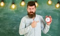 Teacher in eyeglasses holds alarm clock. Lateness concept. Man with beard and mustache on strict face pointing at clock