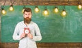 Teacher in eyeglasses holds alarm clock. Schedule and regime concept. Bearded hipster holds clock, chalkboard on