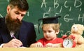 Teacher in formal wear and pupil in mortarboard in classroom, chalkboard on background. Father checking hometask, helps Royalty Free Stock Photo