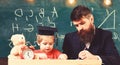 Teacher in formal wear and pupil in mortarboard in classroom, chalkboard on background. Father checking homework, helps Royalty Free Stock Photo