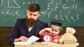 Teacher in formal wear and pupil in mortarboard in classroom, chalkboard on background. Teacher, father checking Royalty Free Stock Photo