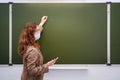 A teacher in a medical mask with a phone in hand writes a text on a school blackboard, copy space. School quarantine education