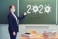 A teacher is standing in a medical mask at the blackboard with chalk 2020, copy space. Woman school teacher with books at desk Royalty Free Stock Photo