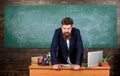 Teacher strict serious bearded man lean on table chalkboard background. Teacher looks threatening. School principal