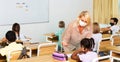 Teacher woman in protective mask helping schoolkid during lesson