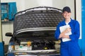 Female mechanic in blue coveralls checking open car hood in workshop holding clipboard, copy space Royalty Free Stock Photo