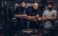 Team of professional bearded cooks dressed in uniforms posing with knives in kitchen.