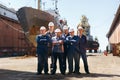 Team of shipyard workers stand proudly at dry dock. Marine engineers in helmets, repair crew with vessel. Dockyard Royalty Free Stock Photo