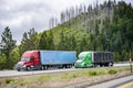 Team of two big rig semi trucks with semi trailers carry cargo driving on the highway road along the huge mountain with thin semi- Royalty Free Stock Photo