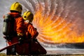 Team of two firefighters wearing full uniform with oxygen tank on back holding pipe spread water and face to wide huge orange hot