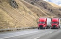 Team of two red big rigs semi trucks transporting cargo in dry van semi trailers running together on the multiline mountain Royalty Free Stock Photo