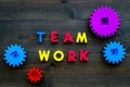 Teamwork concept. Text teamwork lined with colored letters near toy gears on dark wooden background top view copy space